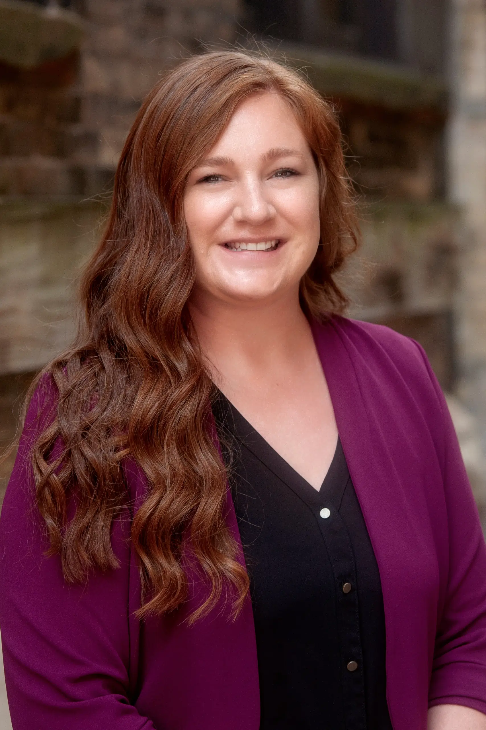 Portrait of a smiling woman with long wavy auburn hair wearing a purple blazer and black blouse.
