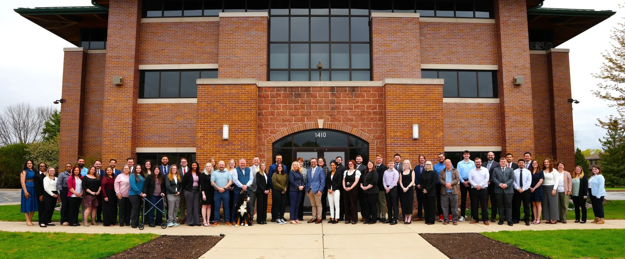 Large diverse group of professionally dressed people standing outside brick office building with glass windows and entrance door numbered 1410.