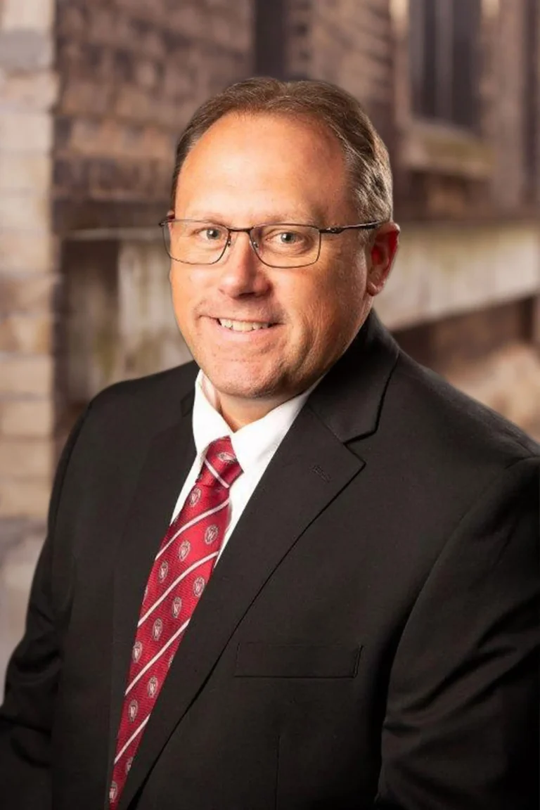 Professional middle-aged man wearing glasses, a black suit, white shirt, and red patterned tie.