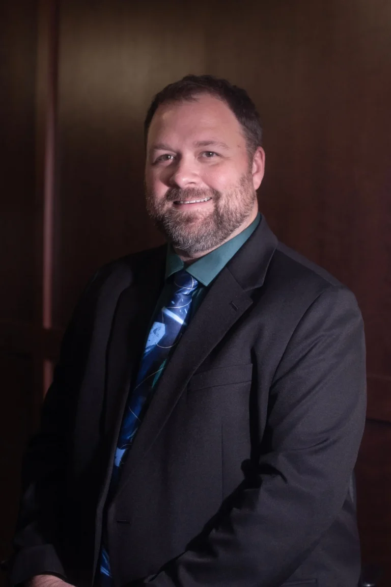 Professional portrait of a smiling man with beard wearing a dark suit and blue patterned tie.