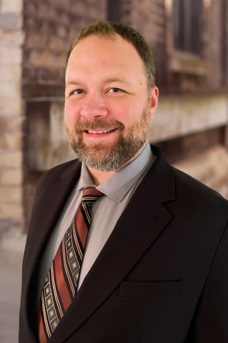 Portrait of a smiling man with a beard wearing a dark suit and patterned tie.
