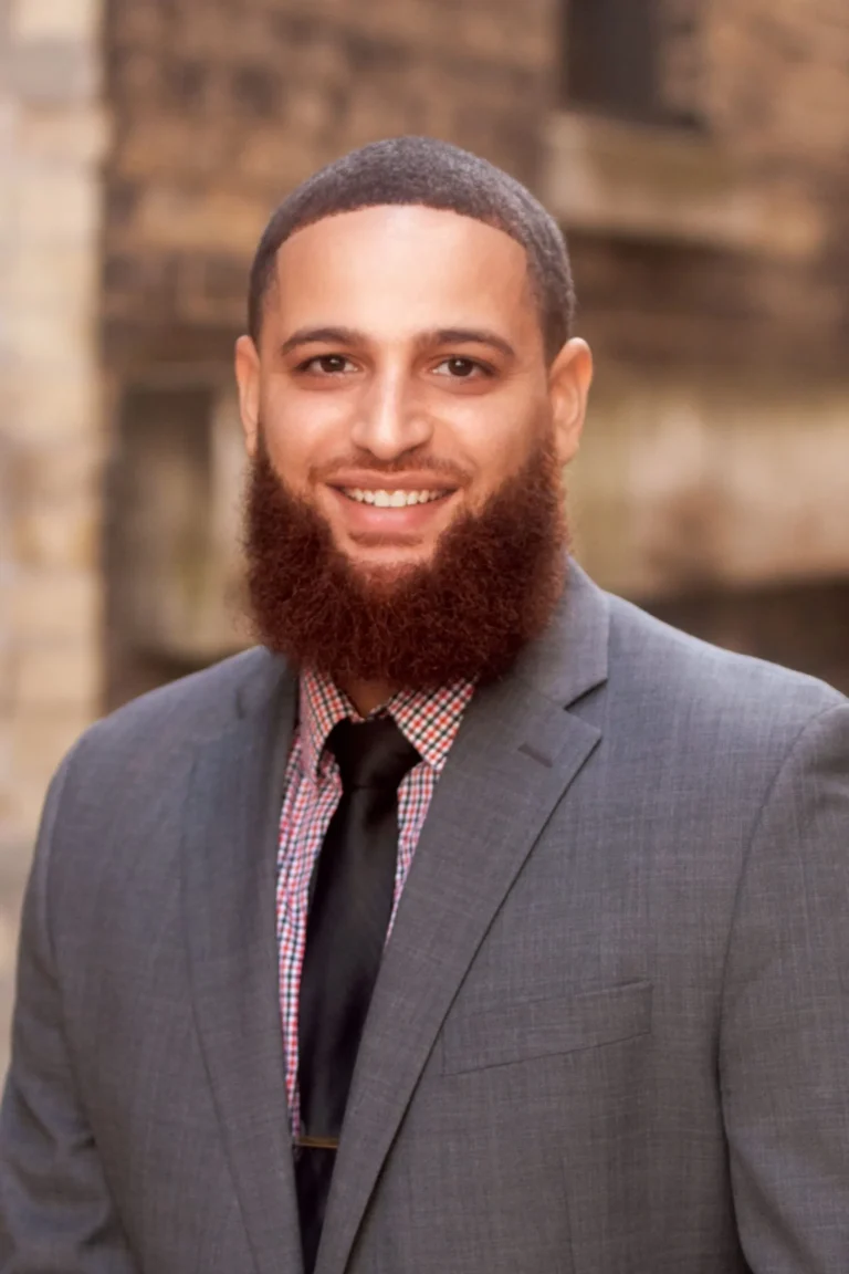 Professional portrait of a smiling man with a full beard, wearing a gray suit and black tie.