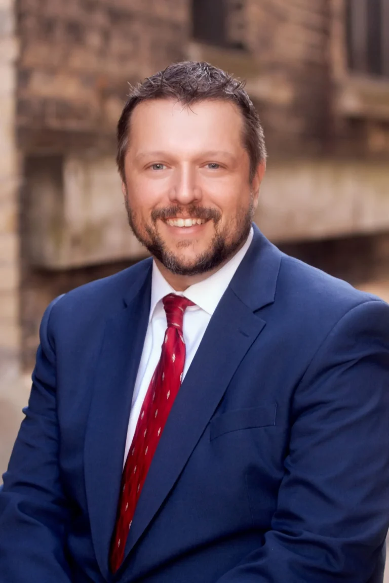 Portrait of a smiling man in a navy blue suit and red tie against a blurred brick background.