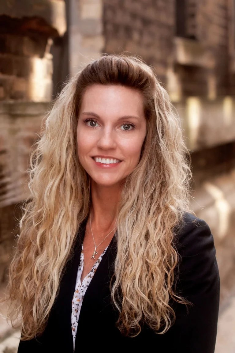 Professional headshot of a smiling woman with long blonde curly hair, wearing a black blazer.