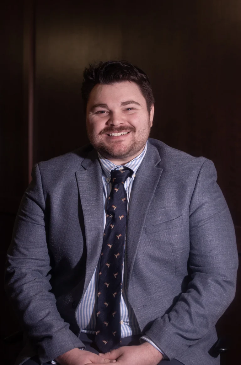 Portrait of a smiling man wearing a gray suit, striped shirt, and patterned tie against a dark background.