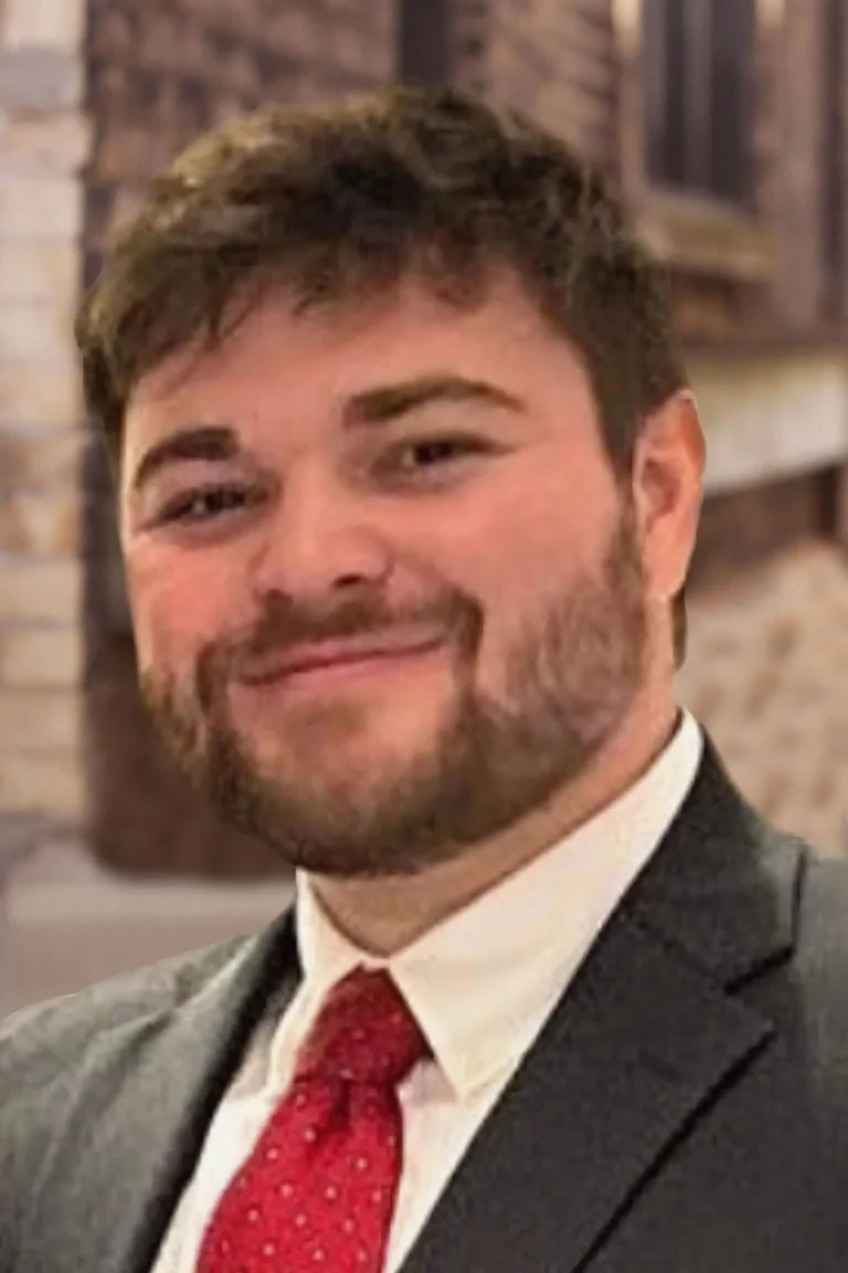 Portrait of a smiling man with beard wearing a dark suit and red tie against a blurred brick background.