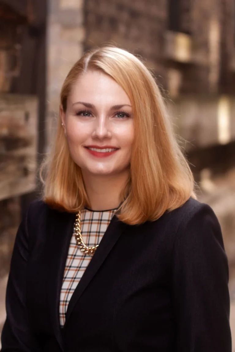 Professional portrait of a smiling woman with blonde hair, wearing a black blazer and gold chain necklace.