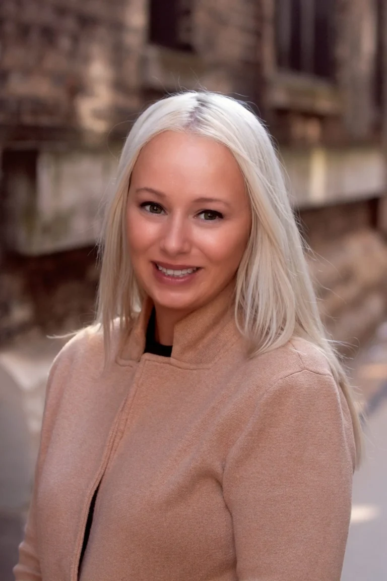 Professional portrait of a smiling woman with long blonde hair wearing a tan coat.