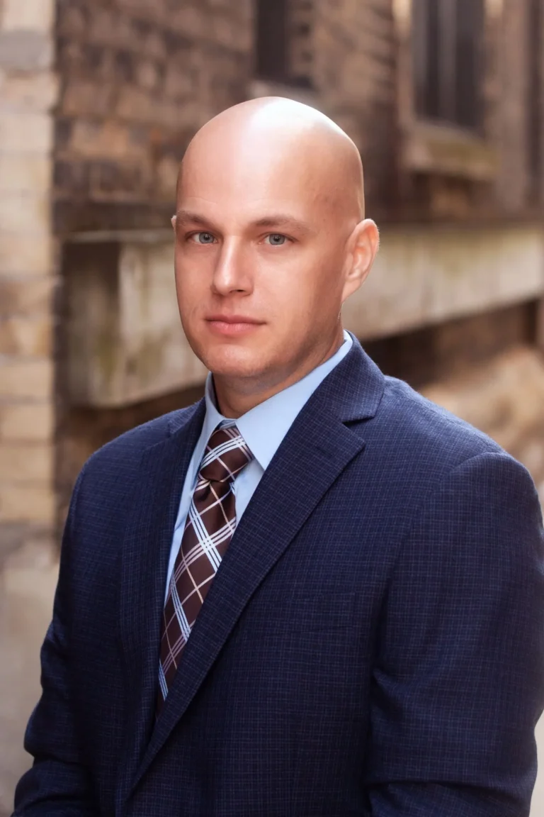 Professional portrait of a bald man in a blue suit and checkered tie against a blurred brick background.