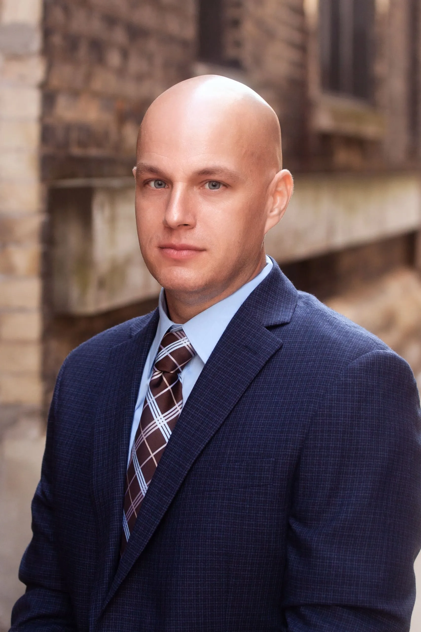 Professional portrait of a bald man in a blue suit and checkered tie against a blurred brick background.