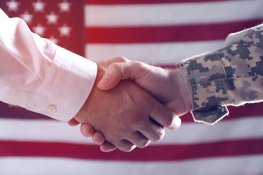 Handshake between a person in business attire and a soldier in camouflage uniform, American flag background.