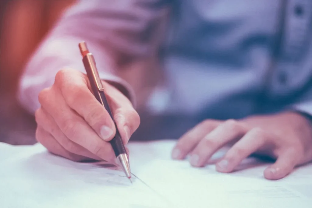 Close-up of a person writing with a black pen on white paper, wearing a blue shirt.