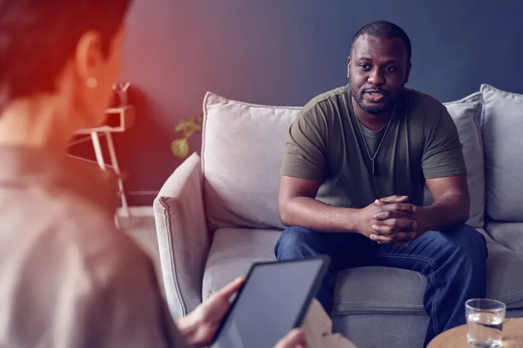 Man in casual clothing seated on a gray sofa, speaking to a person holding a tablet.