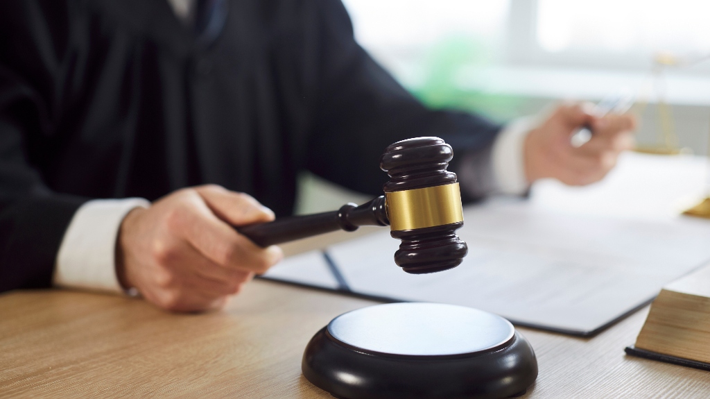 Judge in black robe holding wooden gavel above sound block on desk.