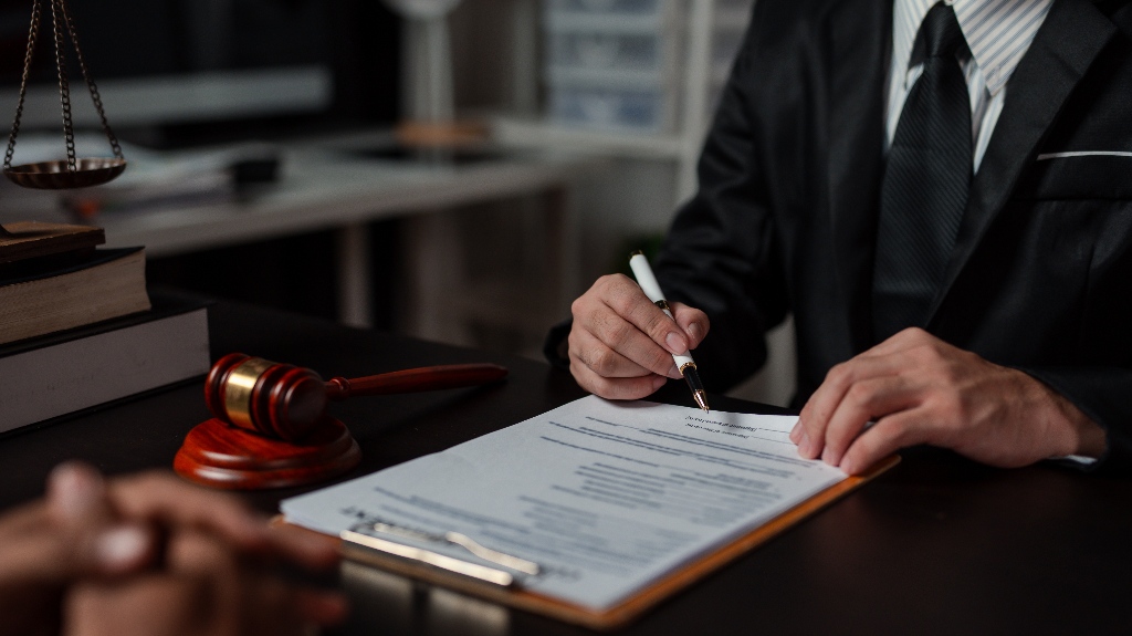 Person in a dark suit signing legal documents at a desk with gavel and scales of justice.