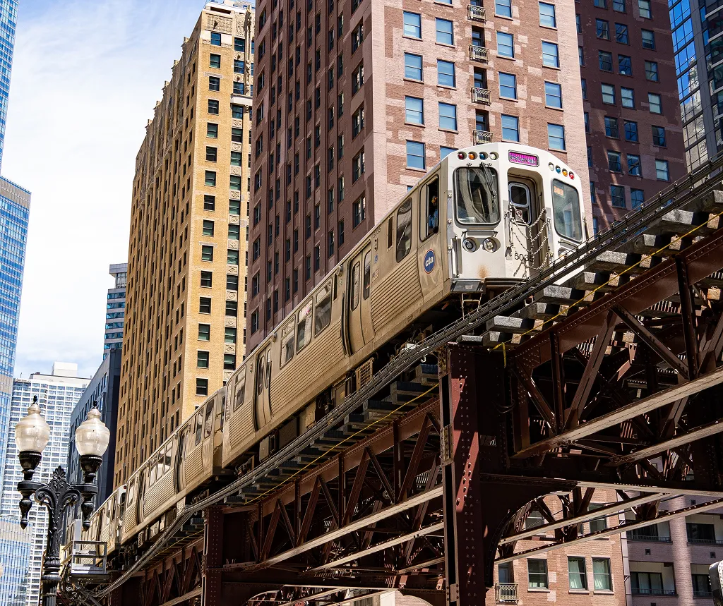 Elevated silver train traveling on rust-colored steel tracks through urban cityscape with tall buildings.