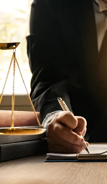 Person in dark suit writing on paper beside golden balance scale and law books.