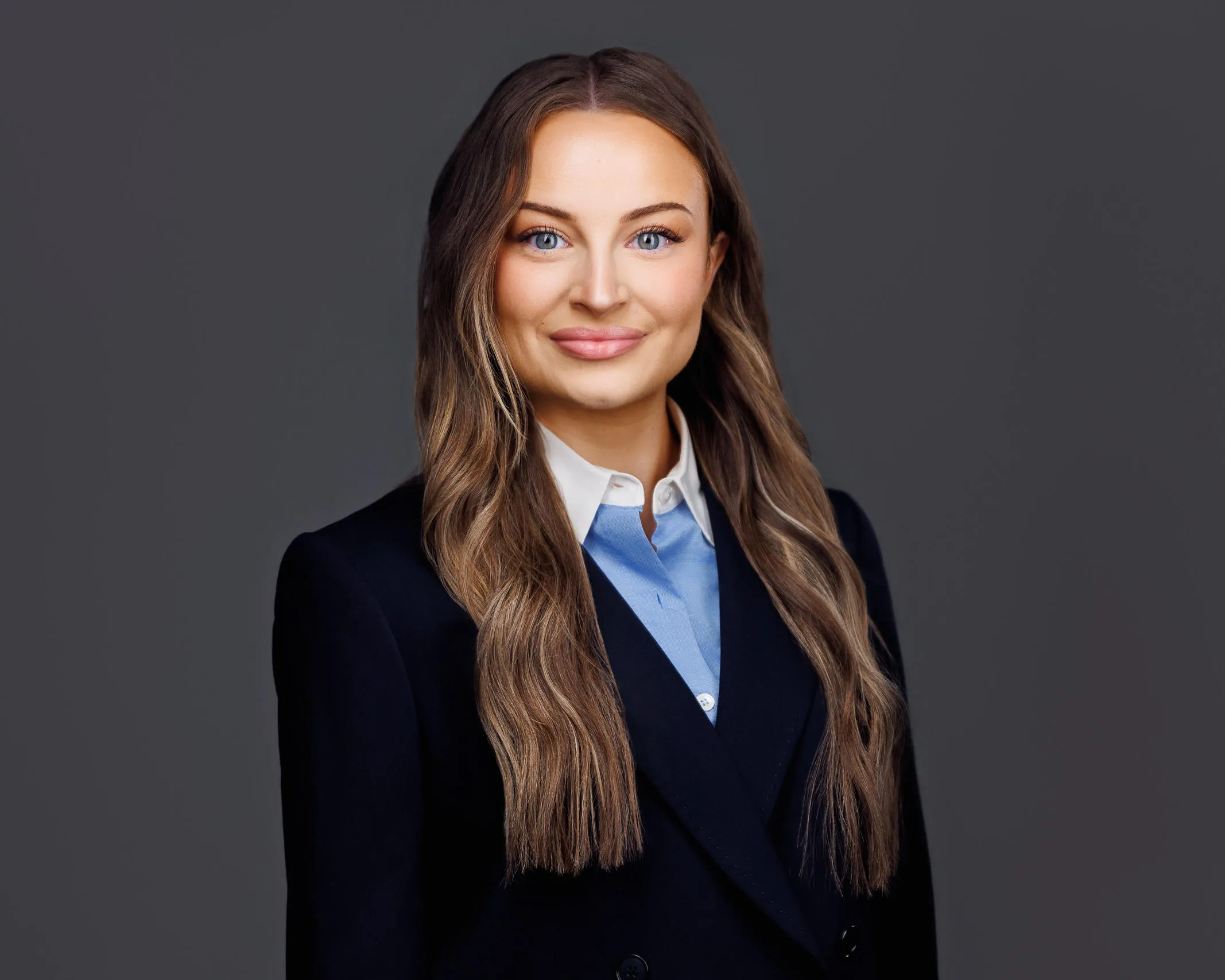 Professional headshot of a woman with long brown hair, blue eyes, wearing a dark blazer and light blue collared shirt.
