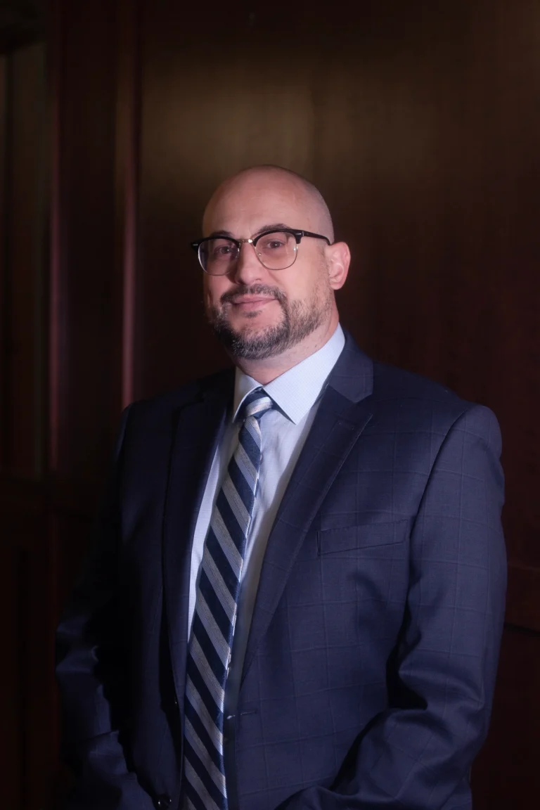 Professional portrait of a bald man with glasses wearing a navy suit and striped tie.