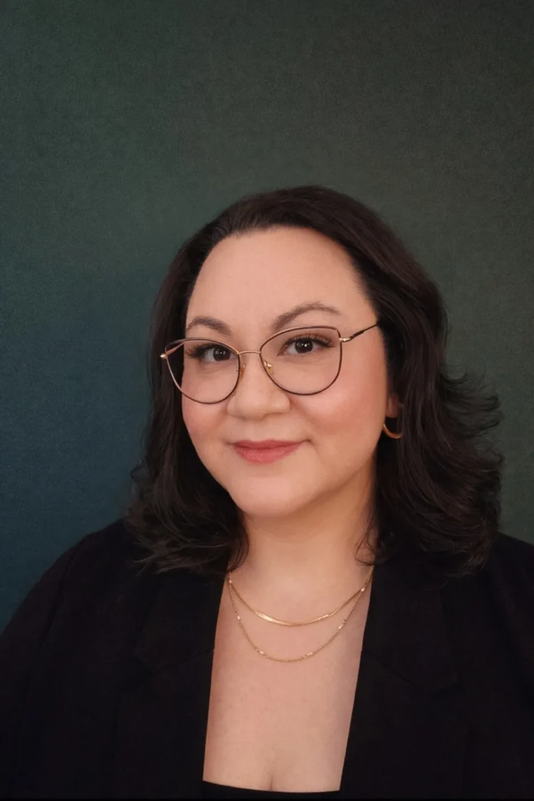 Professional portrait of a woman with dark hair, glasses, gold jewelry, and a dark blazer.
