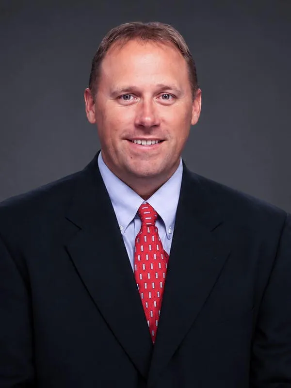 Professional headshot of a smiling man wearing a black suit, light blue shirt, and red patterned tie.