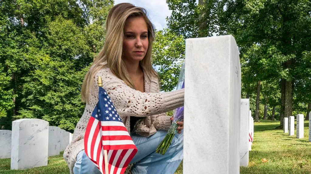 Young woman placing flowers at a white marble gravestone in a cemetery with American flags.