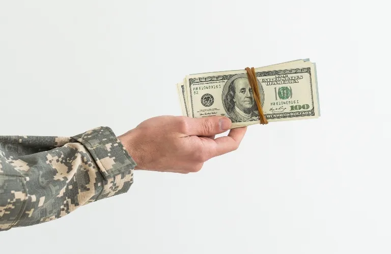 Hand in camouflage military uniform holding a stack of US hundred-dollar bills secured with a rubber band.