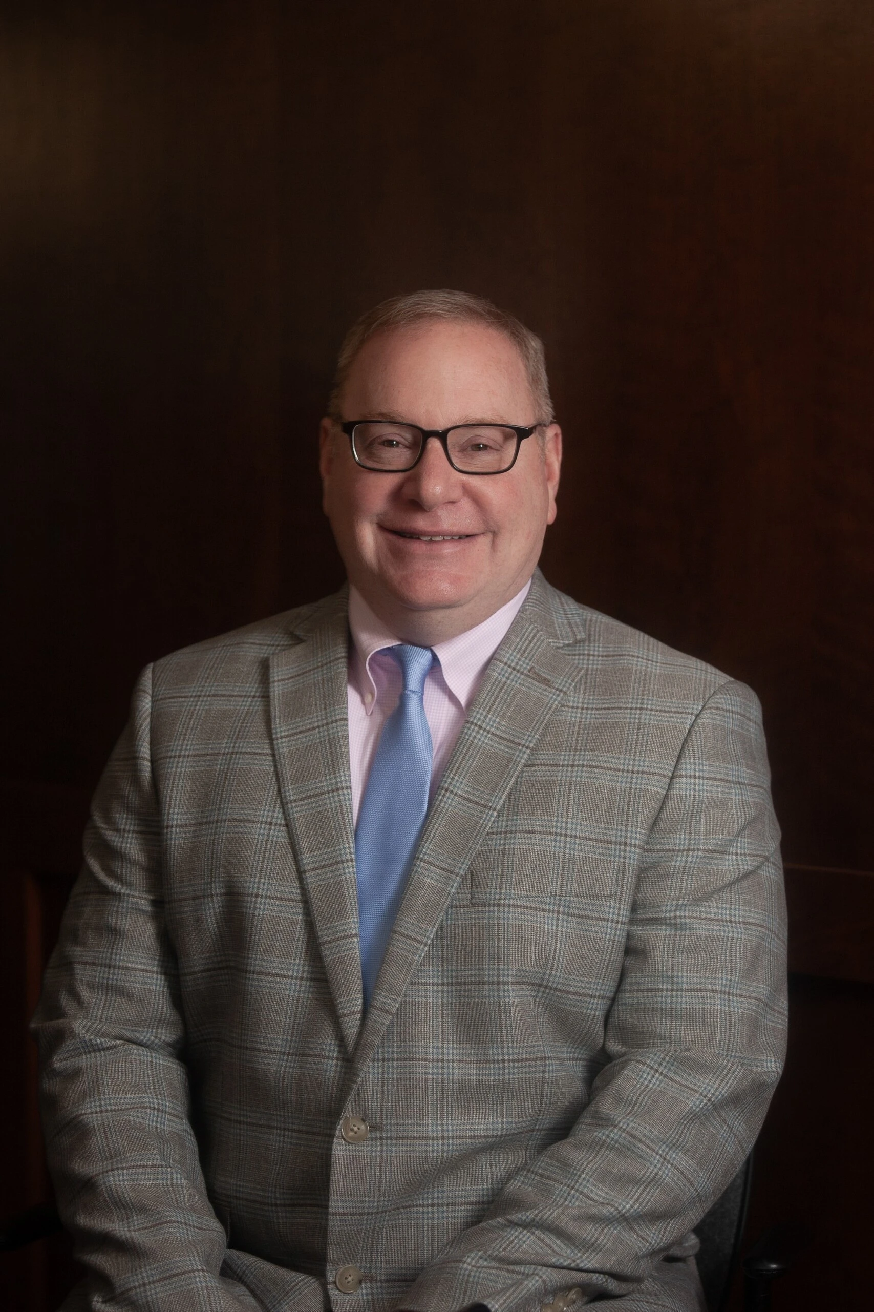 Middle-aged man wearing glasses, gray plaid suit, light blue tie, smiling against dark background.