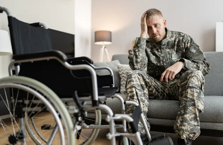 A male soldier in camouflage uniform sitting on a gray sofa, looking distressed, with an empty wheelchair in the foreground.