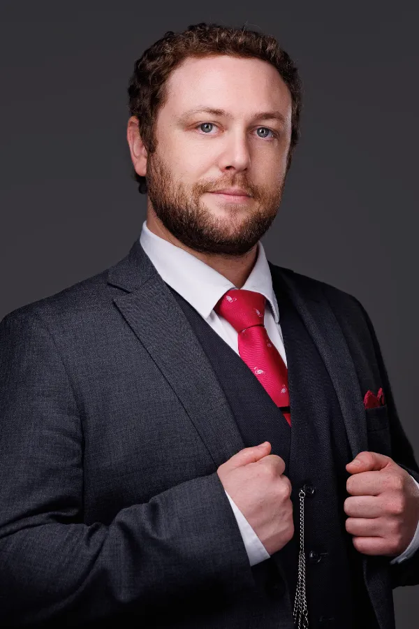 Professional man with curly hair and beard wearing dark gray suit and red tie against gray background.
