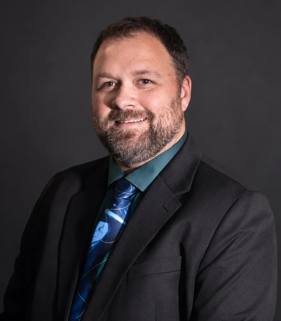 Professional headshot of a smiling bearded man wearing a black suit and blue patterned tie.