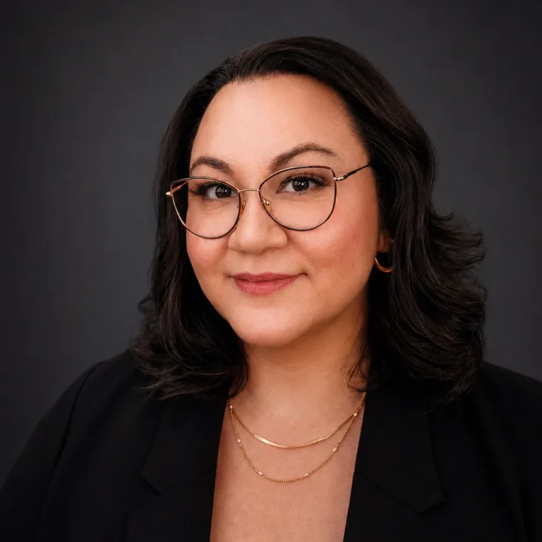 Professional headshot of a woman with dark hair, glasses, and gold jewelry against a dark background.