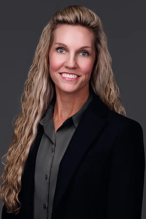 Professional portrait of a smiling woman with long blonde hair wearing a dark blazer and gray shirt.