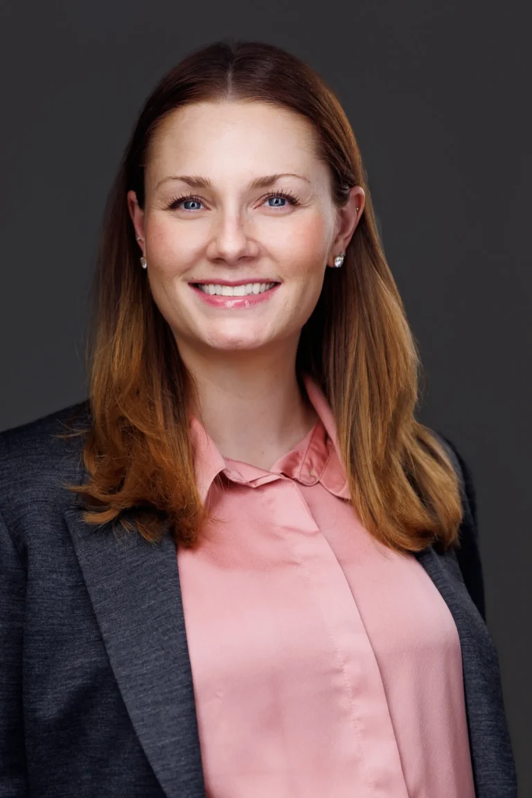 Professional headshot of a smiling woman with long brown hair, wearing a pink blouse and dark blazer.