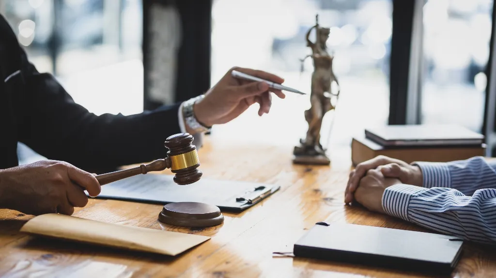 Two people at a wooden table with a judge's gavel, legal books, and a Lady Justice statue.