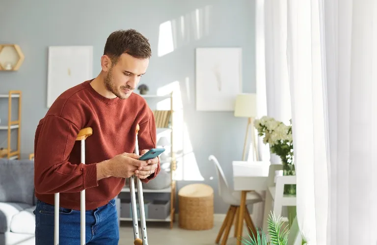Young man in a red sweater using crutches and looking at smartphone in a bright living room.