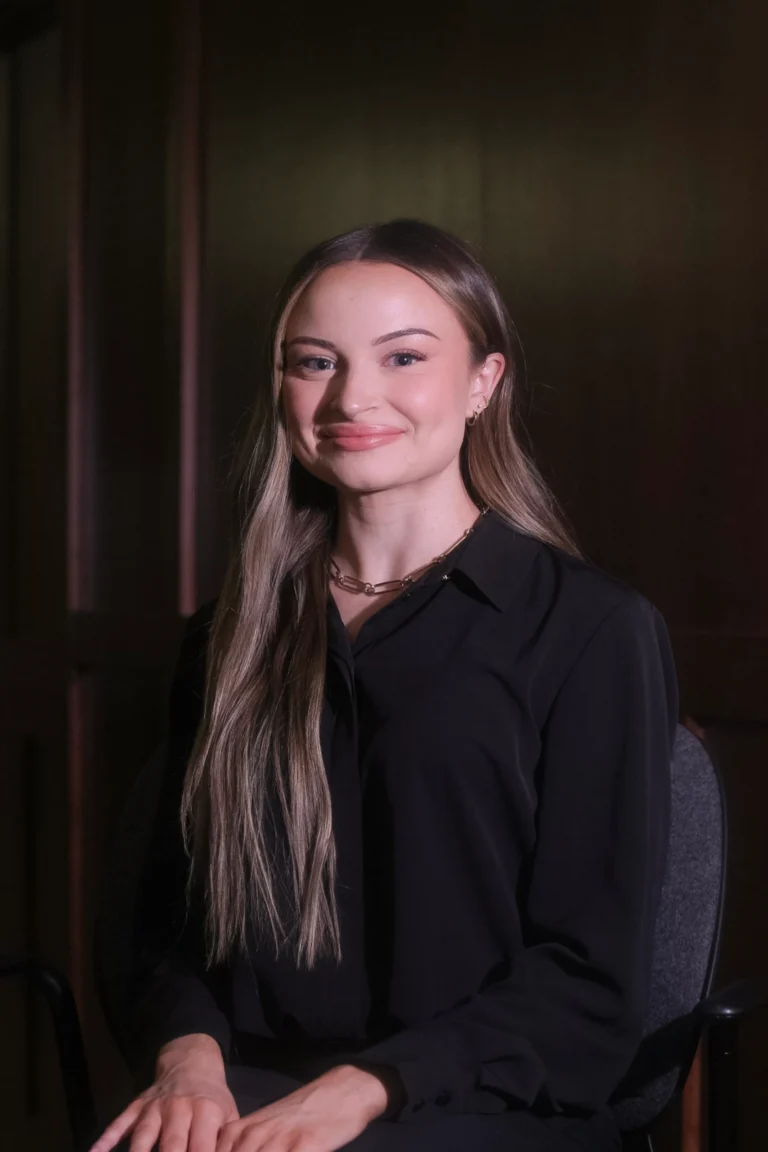 Portrait of a young woman with long hair, wearing a black blouse and gold necklace, seated indoors.