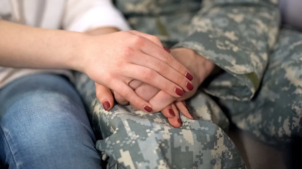 Close-up of two people holding hands, one wearing military camouflage uniform and the other in casual clothing with red nail polish.