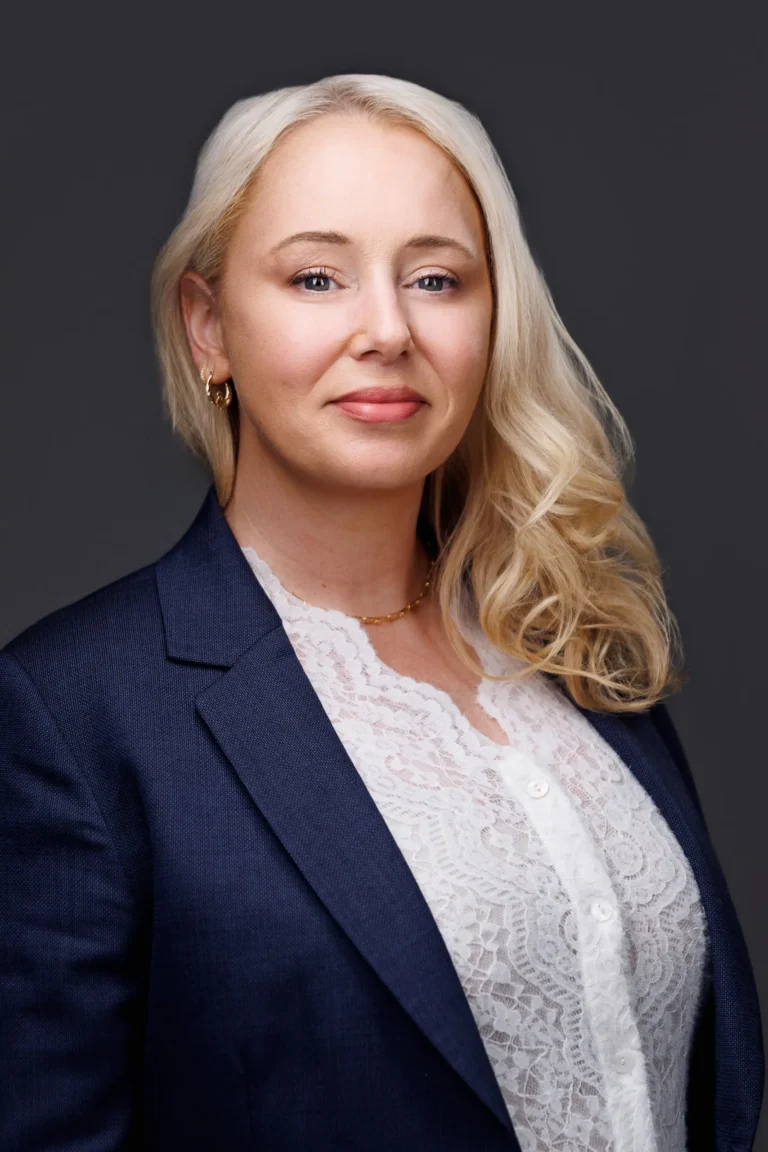 Professional headshot of a blonde woman with curled hair, wearing a navy blazer and white lace blouse.