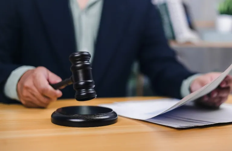 Person in dark suit holding wooden gavel above sound block and reviewing documents on wooden table.