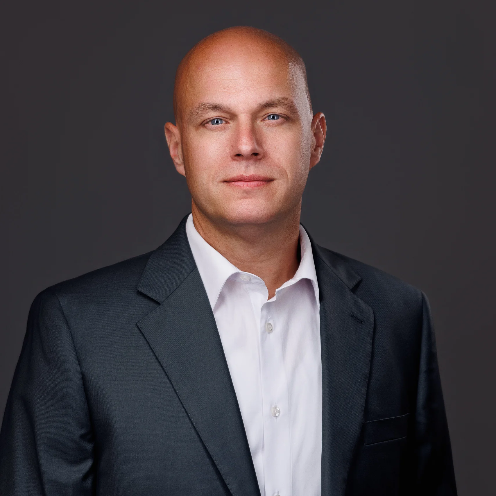 Professional headshot of a bald man with blue eyes wearing a dark suit and white shirt.
