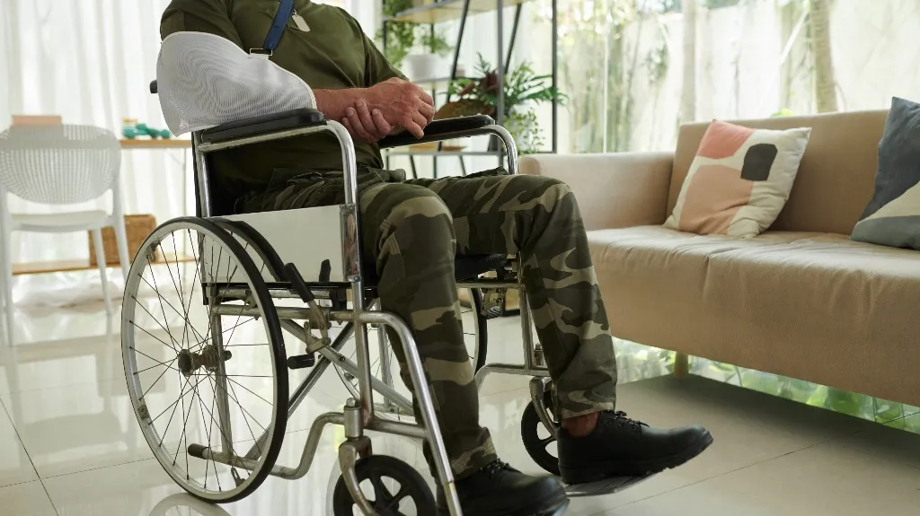 Man in camouflage pants with arm in sling seated in wheelchair in bright living room.