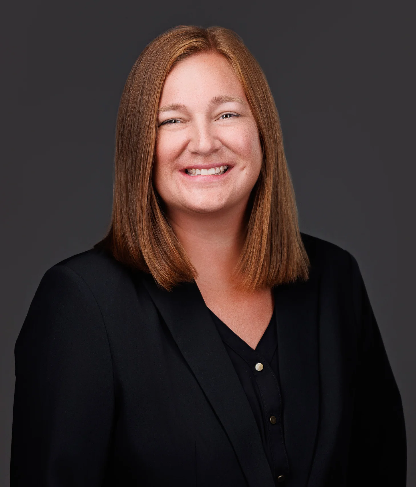 Professional headshot of a smiling woman with straight red hair, wearing a black blazer.