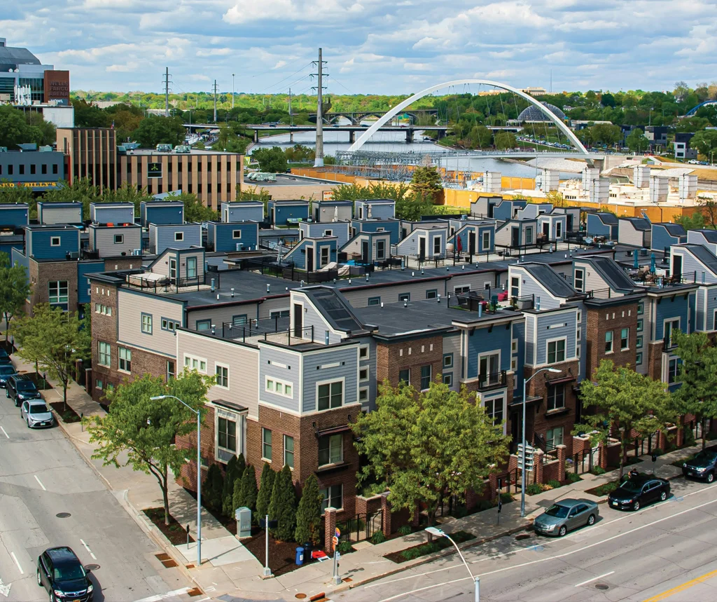 Aerial view of modern residential townhouses with rooftop patios, adjacent streets, trees, and a river bridge in the background.