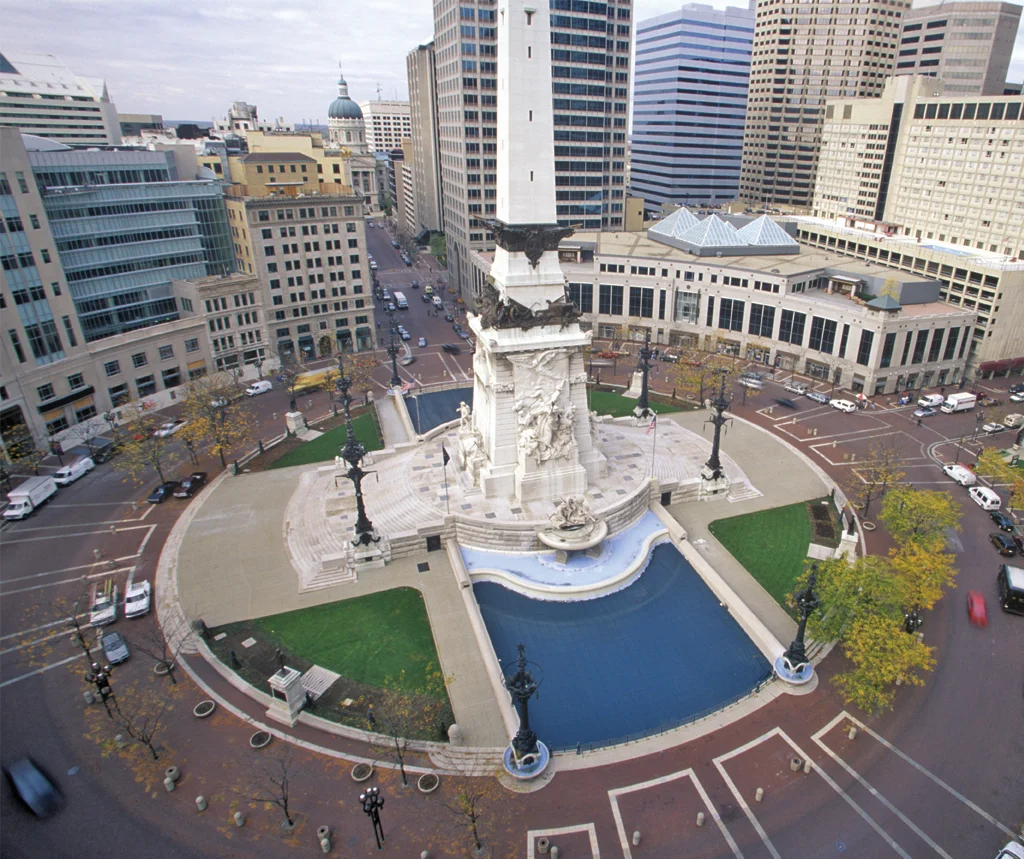 Aerial view of a tall white monument with sculptures, surrounding fountains, and city buildings.