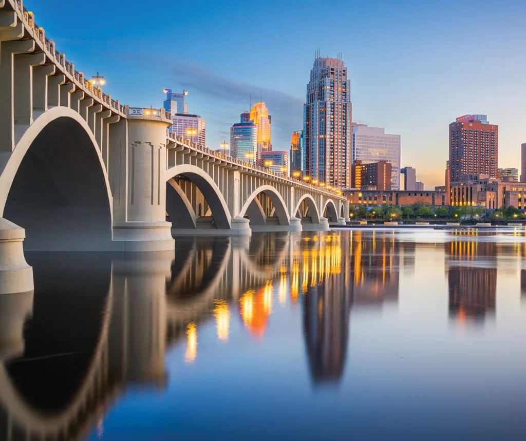 Arched bridge over calm river reflecting city skyline at dusk with illuminated buildings.