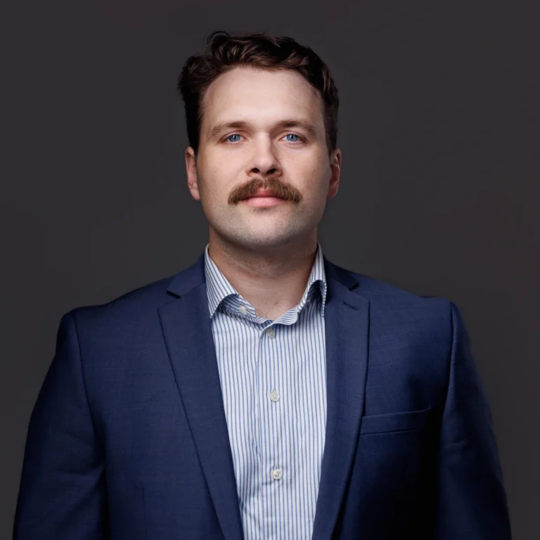 Professional portrait of a man with blue eyes, brown mustache, wearing a blue suit and striped shirt.