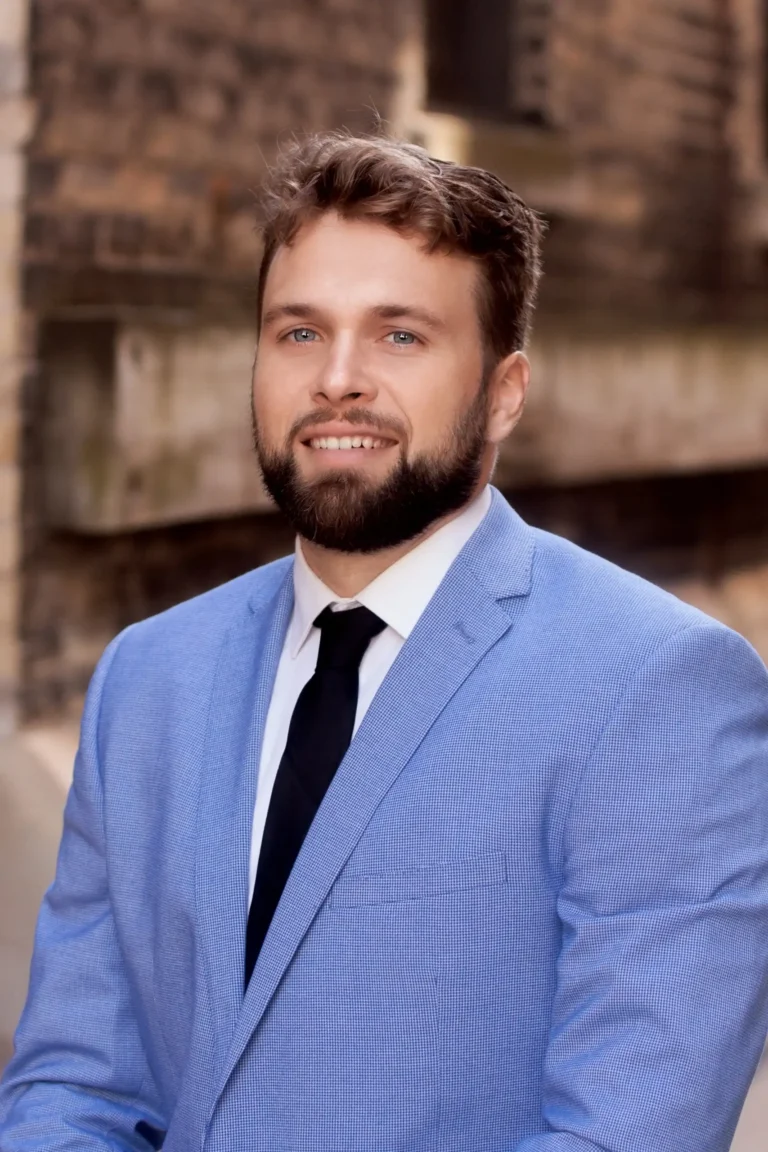 Professional portrait of a smiling man with beard wearing light blue suit and black tie.