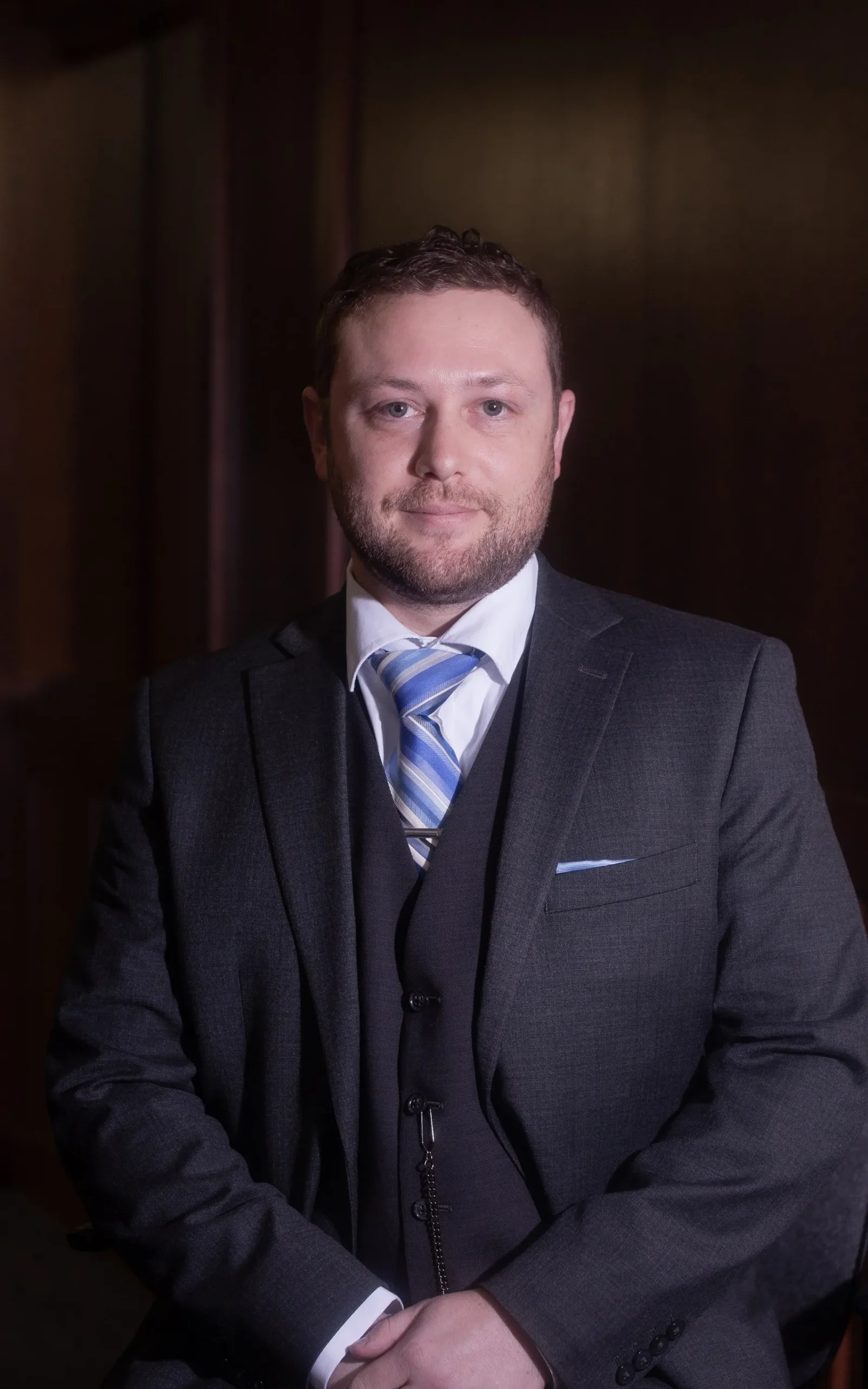Portrait of a man in a dark suit, white shirt, and blue striped tie against a dark background.