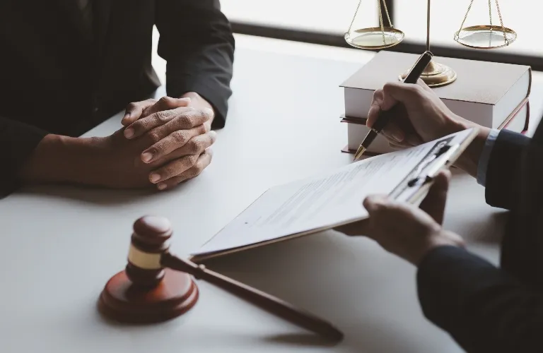 Two individuals at a desk with legal books, scales of justice, gavel, and a document on a clipboard.
